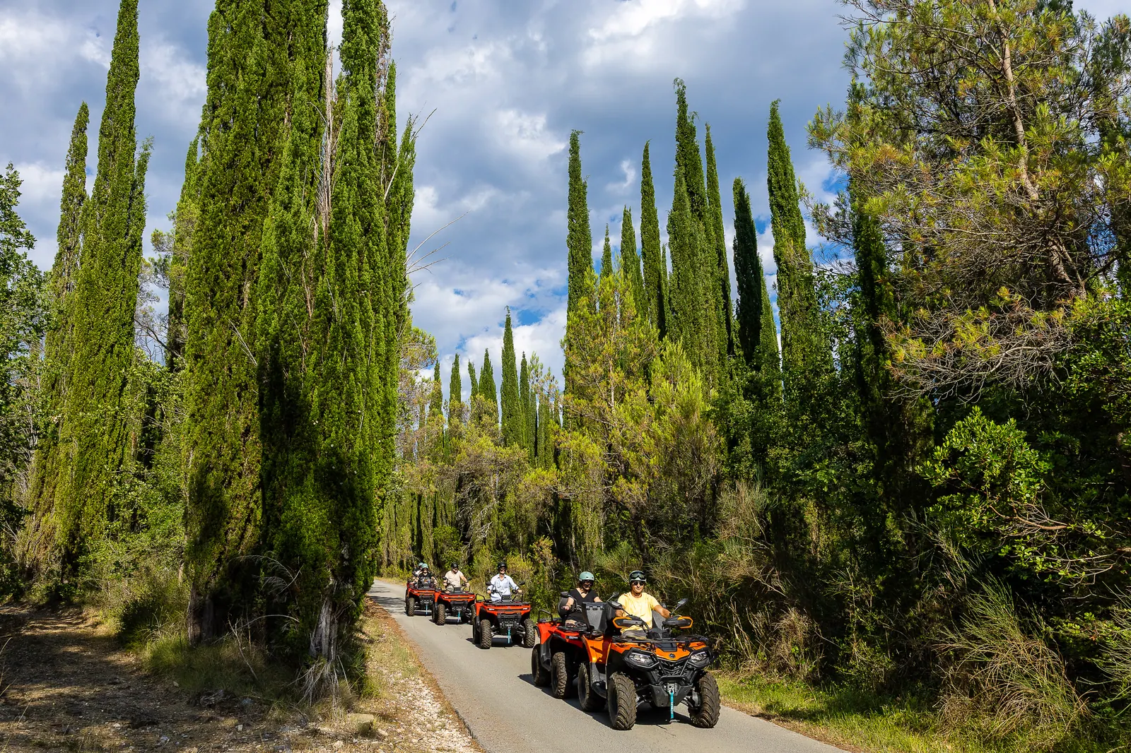 ATV convoy on a road lined with tall cypresses under summer clouds